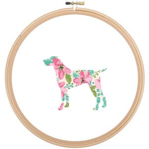 May include: A floral pattern dog silhouette cross-stitch design in a wooden embroidery hoop. The dog is outlined in a light blue and filled with pink and green flowers.