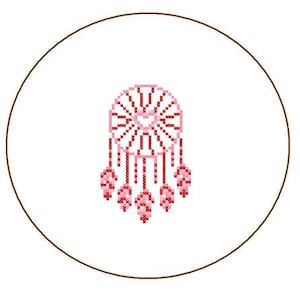May include: A cross-stitch design of a dreamcatcher in a circular frame. The dreamcatcher is primarily pink and red, with a central web design and dangling feather-like embellishments. The frame is a thin brown line on a white background.