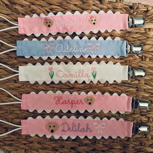 May include: Five personalized pacifier clips with different names and designs. The clips are pink, blue, white, and have a scalloped edge. Each clip features a small embroidered dog, a bow, or a flower.