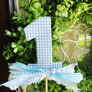 May include: A blue and white gingham fabric number one cake topper with a decorative bow made of blue and white ribbons and lace. The topper is attached to a wooden stick, perfect for a first birthday celebration.