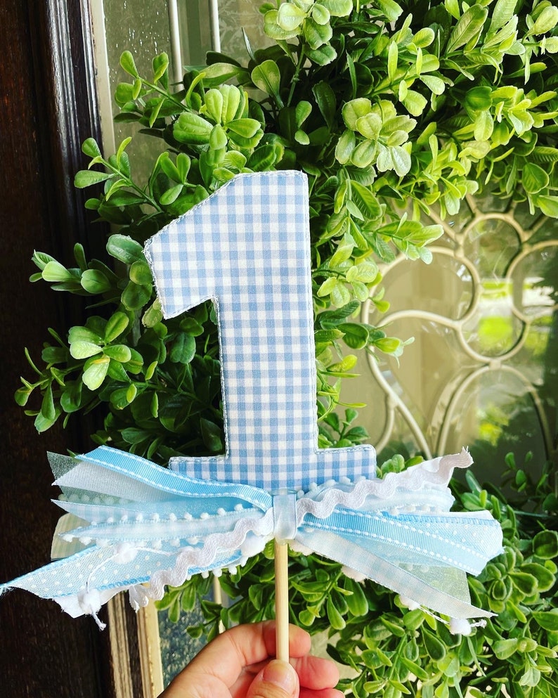 May include: A blue and white gingham fabric number one cake topper with a decorative bow made of blue and white ribbons and lace. The topper is attached to a wooden stick, perfect for a first birthday celebration.