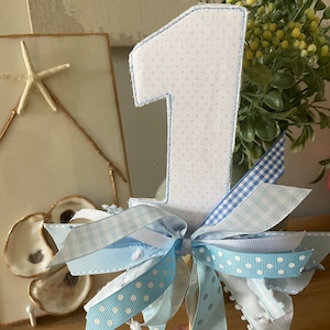 May include: A decorative number one cake topper. The number is white with a blue polka dot pattern and blue stitching. It is adorned with light blue ribbons in various patterns, including polka dots and gingham.
