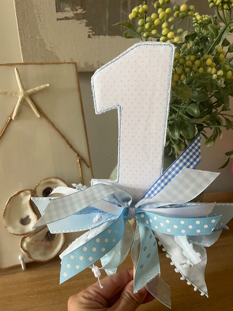 May include: A decorative number one cake topper. The number is white with a blue polka dot pattern and blue stitching. It is adorned with light blue ribbons in various patterns, including polka dots and gingham.