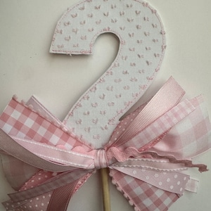 May include: A white fabric number two with pink stitching and a pink bow made of different patterned ribbons.