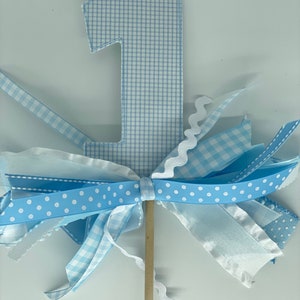 May include: A light blue number one cake topper. The number is made of a gingham pattern fabric. The topper has a bow made of various blue ribbons, including polka dot and gingham patterns. A wooden stick supports the topper.