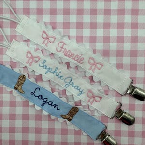May include: Three personalized baby bib clips with embroidered names and designs. The top clip is white with pink bows and the name "Francie". The middle clip is white with pink bows and the name "Sophie Gray". The bottom clip is light blue with brown cowboy boots and the name "Logan".