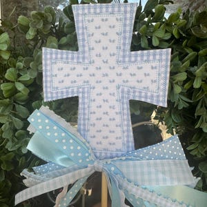 May include: A blue and white checkered cross with a white background and blue embroidered details. The cross is attached to a wooden stick with a blue and white ribbon bow.