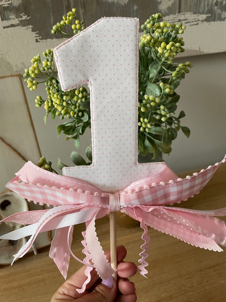 May include: A pink polka-dot fabric number one cake topper with a pink gingham bow and ribbons. The number is outlined in pink stitching and attached to a wooden dowel. Green and yellow floral accents are behind the number.