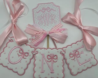 May include: Pink and white banner with the word "ONE" embroidered in pink with a bow on each letter. There is a separate pink and white banner with the letter "S" embroidered in pink with a bow. There are also two pink satin ribbons tied in bows.
