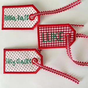 May include: Three red and white fabric gift tags with green embroidered names: Anna Kate, Luke, and Lucy Claire. The tags have a red gingham pattern, red polka dots, and a red scalloped edge.