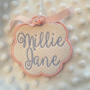 May include: A personalized name tag with the name "Millie Jane" embroidered in blue script on a white fabric with pink polka dots. The tag has a pink scalloped border and is adorned with a pink ribbon.