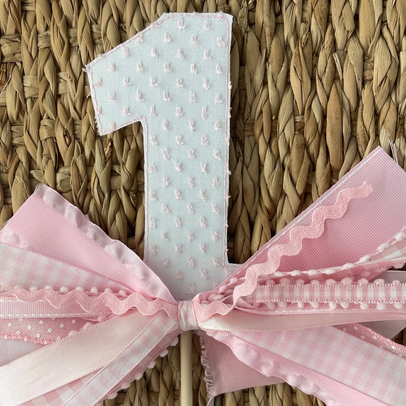 May include: A pink and white number one cake topper with a bow. The number one is white with a textured pattern and pink trim. The bow is made of various pink ribbons with different patterns and textures, including gingham and polka dots.