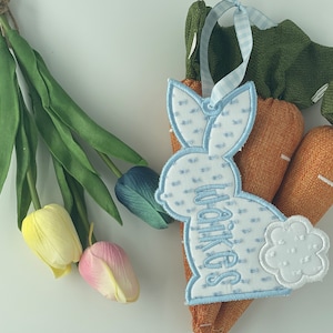 May include: A blue embroidered bunny with the name "Walker's" on it is attached to a brown carrot-shaped fabric ornament. The ornament is decorated with green fabric leaves.
