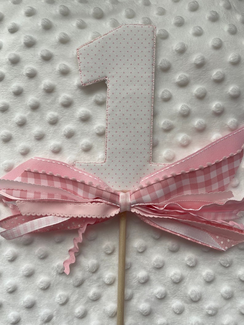 May include: A decorative number one cake topper. The number is white with pink polka dots and a pink ribbon border. A large pink and white gingham bow is attached to the number, and the topper is mounted on a wooden stick.