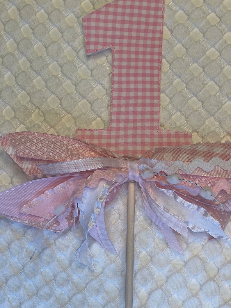 May include: A pink and white gingham number one cake topper with a decorative bow. The bow is made of various pink and white ribbons, including polka dot and ruffled designs. The cake topper is on a wooden stick.