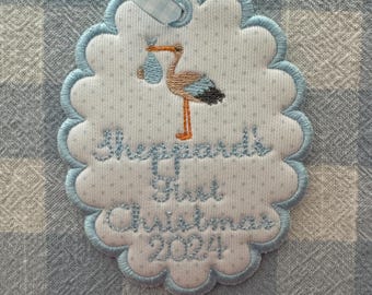 May include: A white and blue scalloped ornament with a stork carrying a baby in a blue bag. The ornament is embroidered with the text "Sheppard's First Christmas 2024".