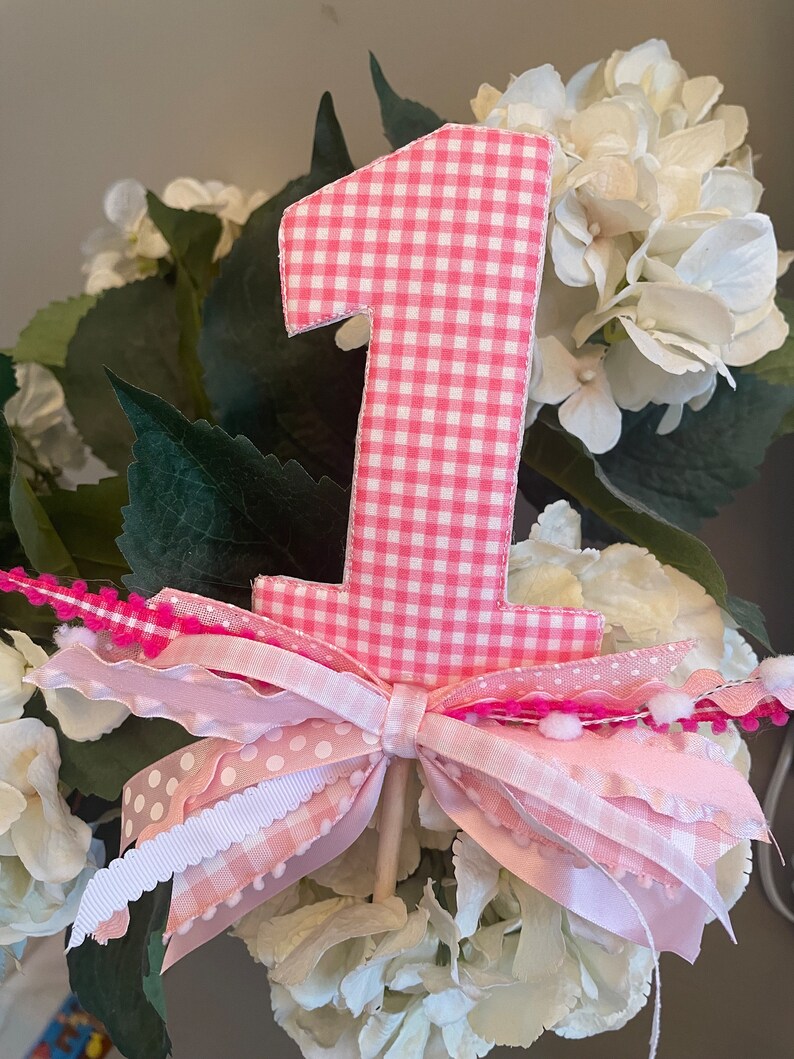 May include: A pink gingham fabric number one cake topper. The number is surrounded by a pink and white ribbon bow and sits atop a wooden dowel. The topper is set against a backdrop of white flowers and green leaves.