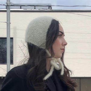 May include: A white and light gray knitted bonnet with tie closures. The bonnet is made of a soft, textured yarn. The person is wearing a black coat.