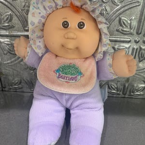 Puede incluir: Una muñeca Cabbage Patch Kids con cara redonda, pelo de lana naranja y un pijama de forro polar morado claro. La muñeca lleva un gorro con volantes con un patrón morado y amarillo y un babero rosa con el texto bordado Cabbage Patch Babyland.