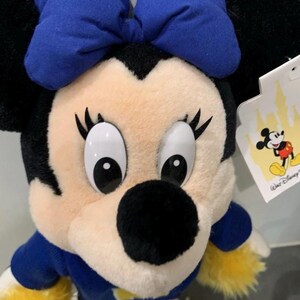 Pu&ograve; includere: Un peluche di Minnie Mouse con un fiocco blu, orecchie nere e un completo blu e bianco. La bambola ha grandi occhi bianchi, ciglia nere e un naso nero. Un'etichetta Disney &egrave; attaccata.