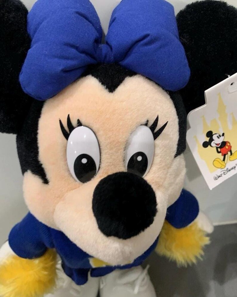 Pu&ograve; includere: Un peluche di Minnie Mouse con un fiocco blu, orecchie nere e un completo blu e bianco. La bambola ha grandi occhi bianchi, ciglia nere e un naso nero. Un'etichetta Disney &egrave; attaccata.