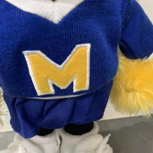 Pu&ograve; includere: Un peluche che indossa una maglia blu e bianca con scollo a V con una grande "M" gialla sul davanti. Il giocattolo ha una gonna a pieghe blu, scarpe da ginnastica bianche e accenti gialli soffici.