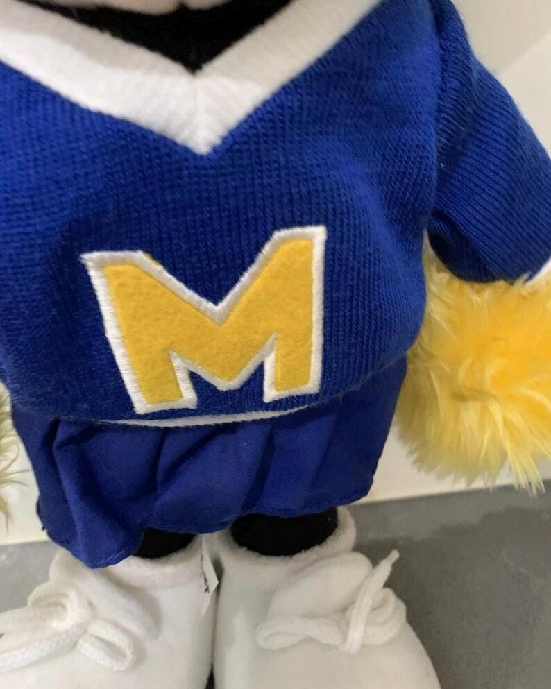 Pu&ograve; includere: Un peluche che indossa una maglia blu e bianca con scollo a V con una grande "M" gialla sul davanti. Il giocattolo ha una gonna a pieghe blu, scarpe da ginnastica bianche e accenti gialli soffici.