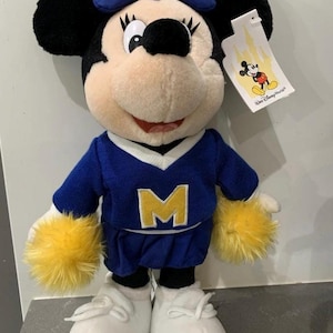 Pu&ograve; includere: Una bambola di peluche di Minnie Mouse con un maglione blu con una "M" gialla, una gonna abbinata e scarpe bianche. La bambola ha orecchie nere, un fiocco blu e pompon gialli sui polsi. &Egrave; attaccato un'etichetta con il logo Walt Disney World.