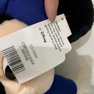 Pu&ograve; includere: Peluche di Minnie Mouse cheerleader, con testa nera, abito blu e dettagli gialli. L'etichetta recita "CHEERLEADER MINNIE" e include un avvertimento per rimuovere l'etichetta prima di dare il giocattolo a un bambino.