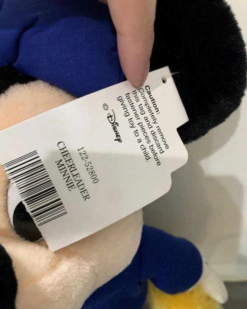 Pu&ograve; includere: Peluche di Minnie Mouse cheerleader, con testa nera, abito blu e dettagli gialli. L'etichetta recita "CHEERLEADER MINNIE" e include un avvertimento per rimuovere l'etichetta prima di dare il giocattolo a un bambino.
