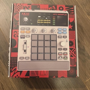 Może przedstawiać: Nowe centrum produkcji muzycznej AKAI Professional MPC SAMPLE, wyświetlane w oryginalnym opakowaniu. Urządzenie jest biało-szare z licznymi przyciskami, pokrętłami i ekranem. Pudełko jest czerwono-czarne z geometrycznymi wzorami.