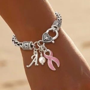 May include: A silver-toned chain link bracelet featuring a heart-shaped toggle clasp, a dangling letter A charm, a single white pearl bead, and a pink ribbon charm. The bracelet has decorative metal spacers with intricate engraved patterns.