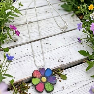 Könnte beinhalten: Eine silberne Halskette mit einem mehrfarbigen Blumenanhänger. Die Blume hat sechs Blütenblätter, jedes mit einem eigenen Muster oder einer eigenen Farbe, darunter Blau, Violett, Grün und Pink, um einen gelben, runden Mittelpunkt.