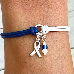 May include: A blue and white leather bracelet with a silver charm in the shape of a ribbon. The charm is attached to a silver ring.