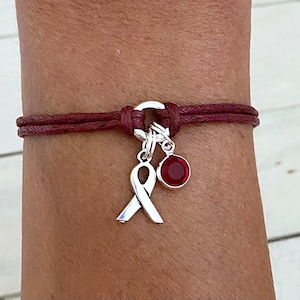May include: A maroon cord bracelet with a silver ribbon charm and a red gemstone charm.
