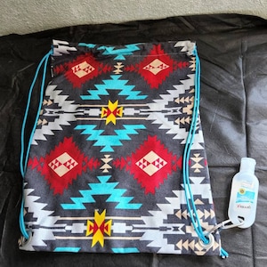 May include: A drawstring bag with a Southwestern geometric pattern in red, turquoise, yellow, and white on a dark gray background. The bag has blue drawstrings and a small bottle of hand sanitizer attached with a carabiner.