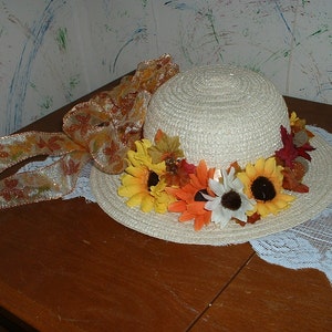 May include: A cream-coloured straw hat decorated with sunflowers, autumn leaves, and a large ribbon. The hat has a wide brim and is adorned with artificial flowers in shades of yellow, orange, and red.