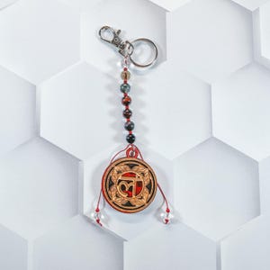 May include: A red and gold charm with the Sanskrit symbol Om, surrounded by a floral design, hangs from a silver key chain with a beaded string of red, black and green stones.