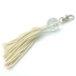 Puede incluir: Llavero con borla de macramé color crema con una cuenta de cristal transparente facetada y un cierre plateado. La borla está hecha de algodón tejido suave. Un accesorio decorativo para llaves o bolsos.