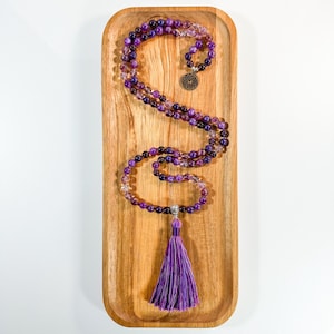 May include: A purple amethyst mala necklace with a tassel and a silver charm. The necklace is displayed on a wooden tray.