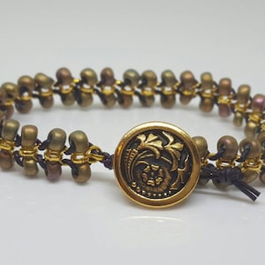 Könnte beinhalten: Ein gold- und braunes Perlenarmband mit einem goldenen Knopfverschluss mit floralem Design. Der Knopf befindet sich in der Mitte des Armbands und hat einen dunkelbraunen Hintergrund.