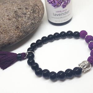 Puede incluir: Pulsera de cuentas negras y moradas con un dije de Buda plateado. La pulsera está hecha con cuentas de lava negra y cuentas moradas.