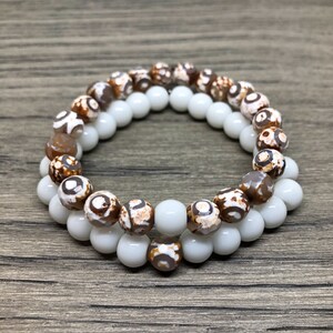 May include: Two bracelets, one white and one brown with white and brown patterns, are stacked on top of each other. The bracelets are made of round beads.