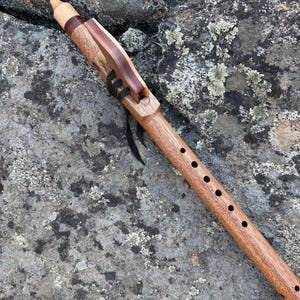 May include: A wooden Native American style flute featuring a light brown body with a darker wood block attached by black leather lacing. The instrument has a smooth finish, a rounded mouthpiece, and six finger holes aligned along the top surface.