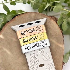 May include: A white Apple Watch band with a floral design. Three metal bands with the text "BLD THNR" and a medical symbol are attached to the watch band. The bands are silver, gold, and rose gold.