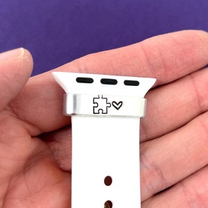 May include: White watch band with a silver metal connector. The connector features a puzzle piece and a heart symbol. The band has several holes for adjustment. The background is a solid purple color.