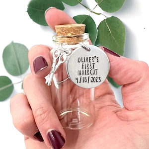 May include: A small glass bottle with a cork top and a silver charm attached. The charm is a round disc with the text "Oliver's First Haircut 4/18/2023" and a pair of scissors.