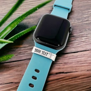May include: A blue Apple Watch with a white band that says "YOUR TEXT" and has a medical symbol on it. The watch is on a wooden surface.
