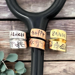 May include: Three metal stethoscope charms with personalized names and designs. One charm is silver with the name "Alexis" and medical symbols. Another is copper with "neppy" and a sun, palm tree, and turtle. The third is gold with "Karen" and dog paw prints.