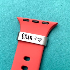 May include: A red silicone Apple Watch band with a silver metal band that says "Erin" with a small airplane icon.